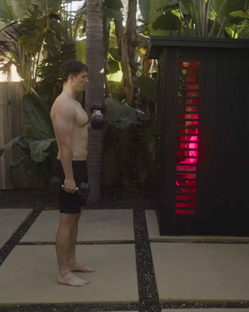 A man standing beside a PEAK outdoor sauna glowing with red light